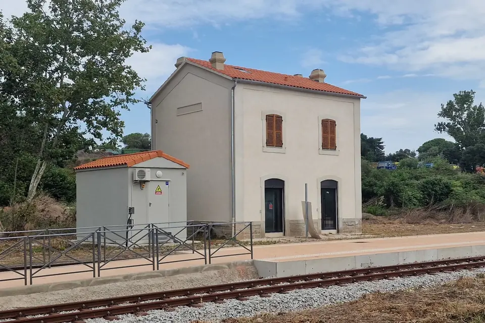 Petite gare de campagne aux murs crème et toit de tuiles, quai et voie ferrée au premier plan, sous un ciel bleu