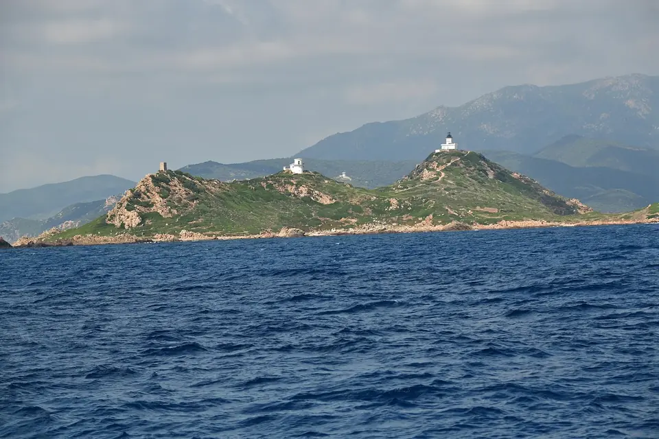 Île rocheuse surmontée d'un phare blanc, mer bleu profond et montagnes de Corse à l'arrière-plan